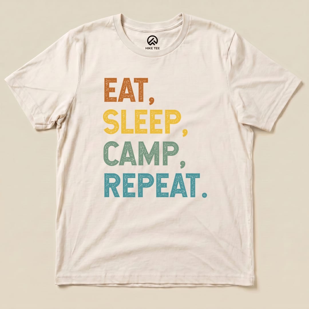 A flat graphic t-shirt featuring the phrase "Eat Sleep Camp Repeat" in colorful, textured lettering.