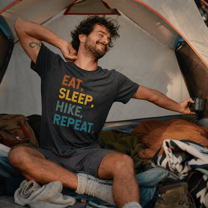 A man is seated in a tent wearing a graphic t-shirt featuring the slogan "Eat, Sleep, Hike, Repeat" in colorful lettering.