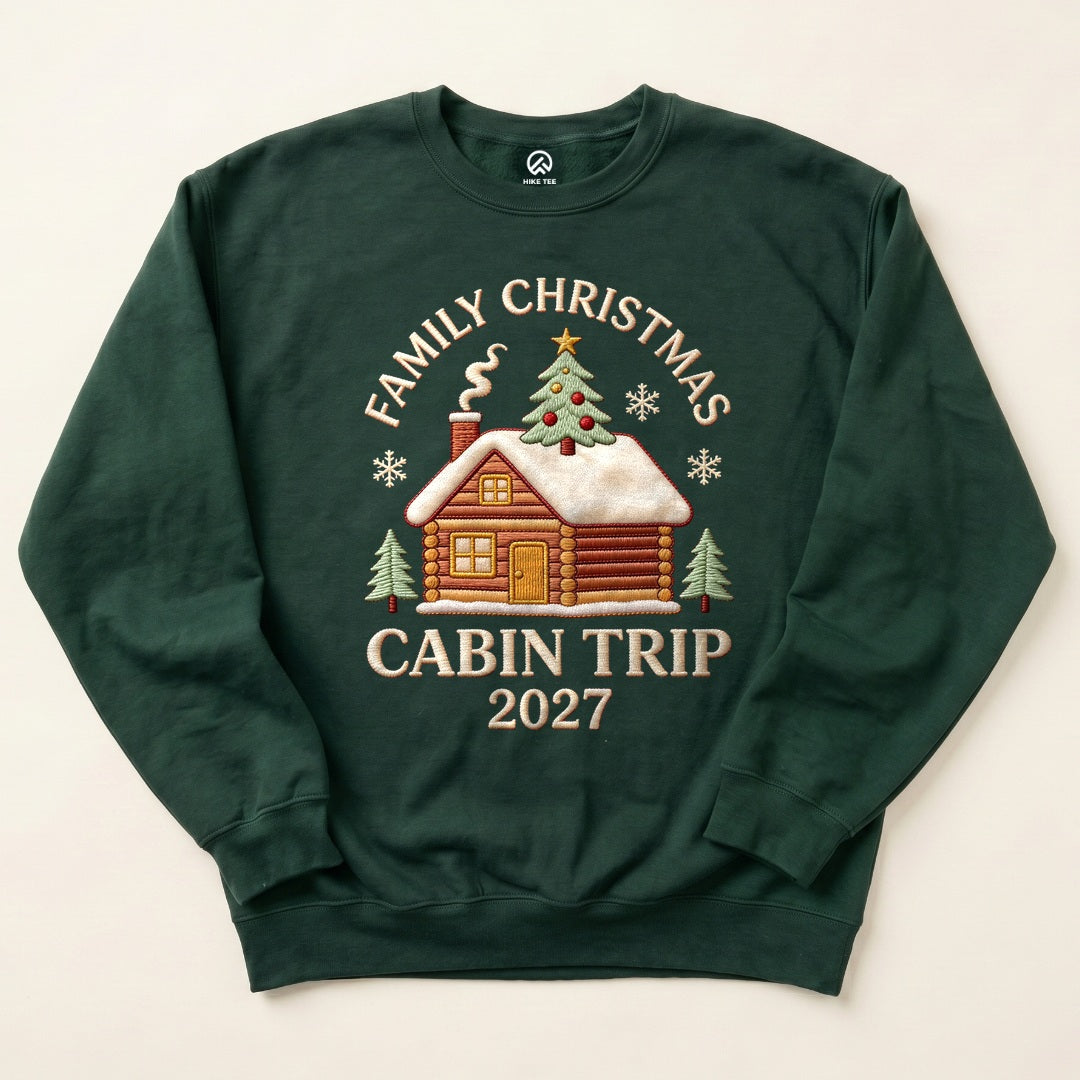 Forest Green Family Christmas Cabin Trip 2027 Sweatshirt with a snowy log cabin and Christmas tree graphic