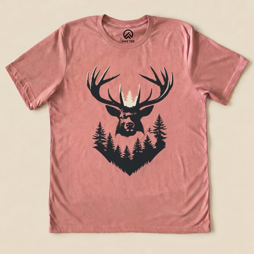 The Elk Shape Shirt features a bold graphic of an elk's head with antlers surrounded by trees, printed on a dusty rose t-shirt.