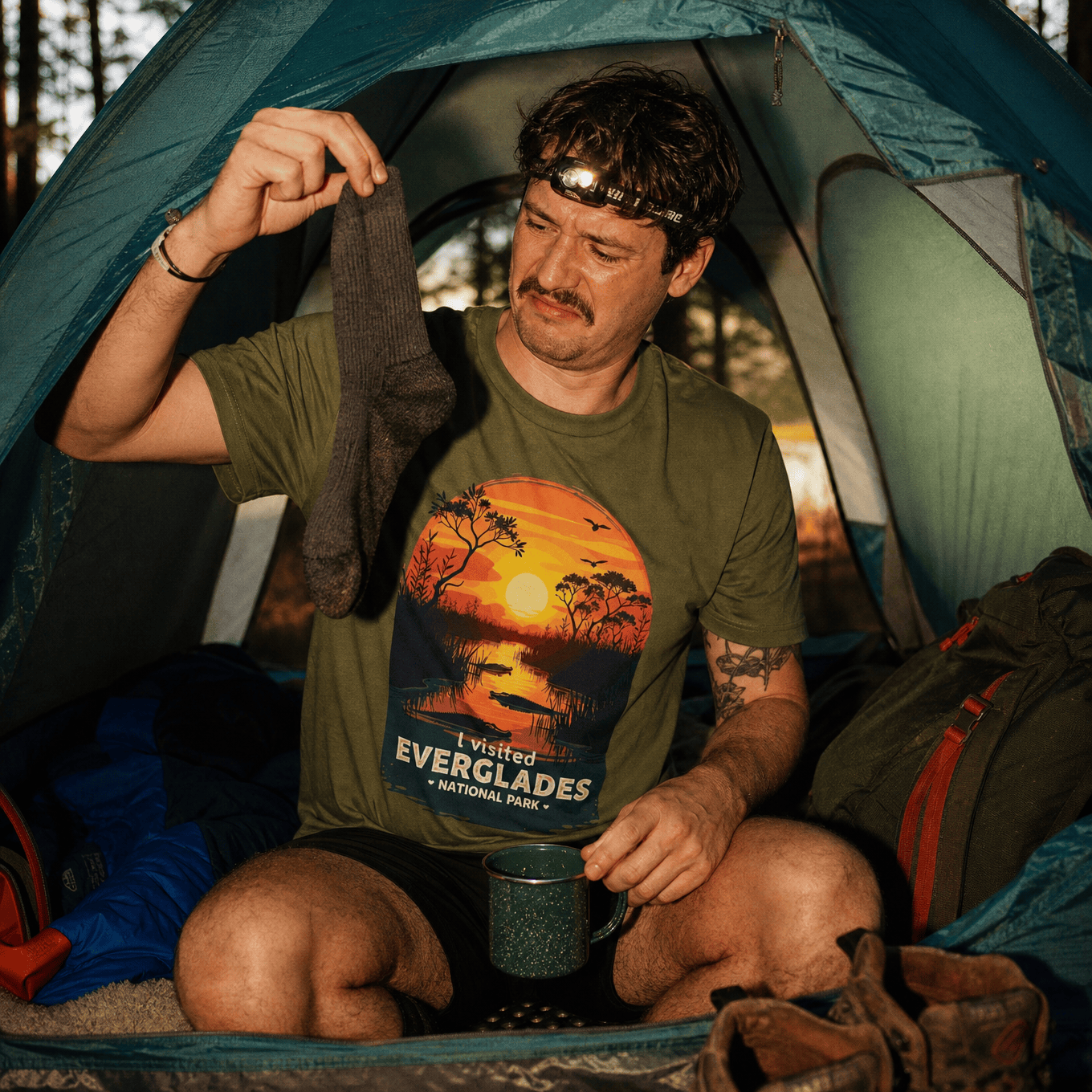 A man inside a tent examines a gray sock while wearing a green graphic t-shirt featuring the text "I Visited Everglades National Park.