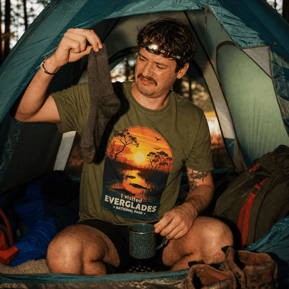A man inside a tent examines a gray sock while wearing a green graphic t-shirt featuring the text "I Visited Everglades National Park.