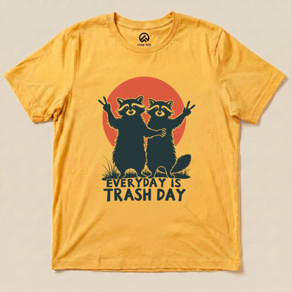 A flat lay of a yellow graphic t-shirt featuring two raccoons and the phrase "Everyday is Trash Day" against a red sun backdrop.