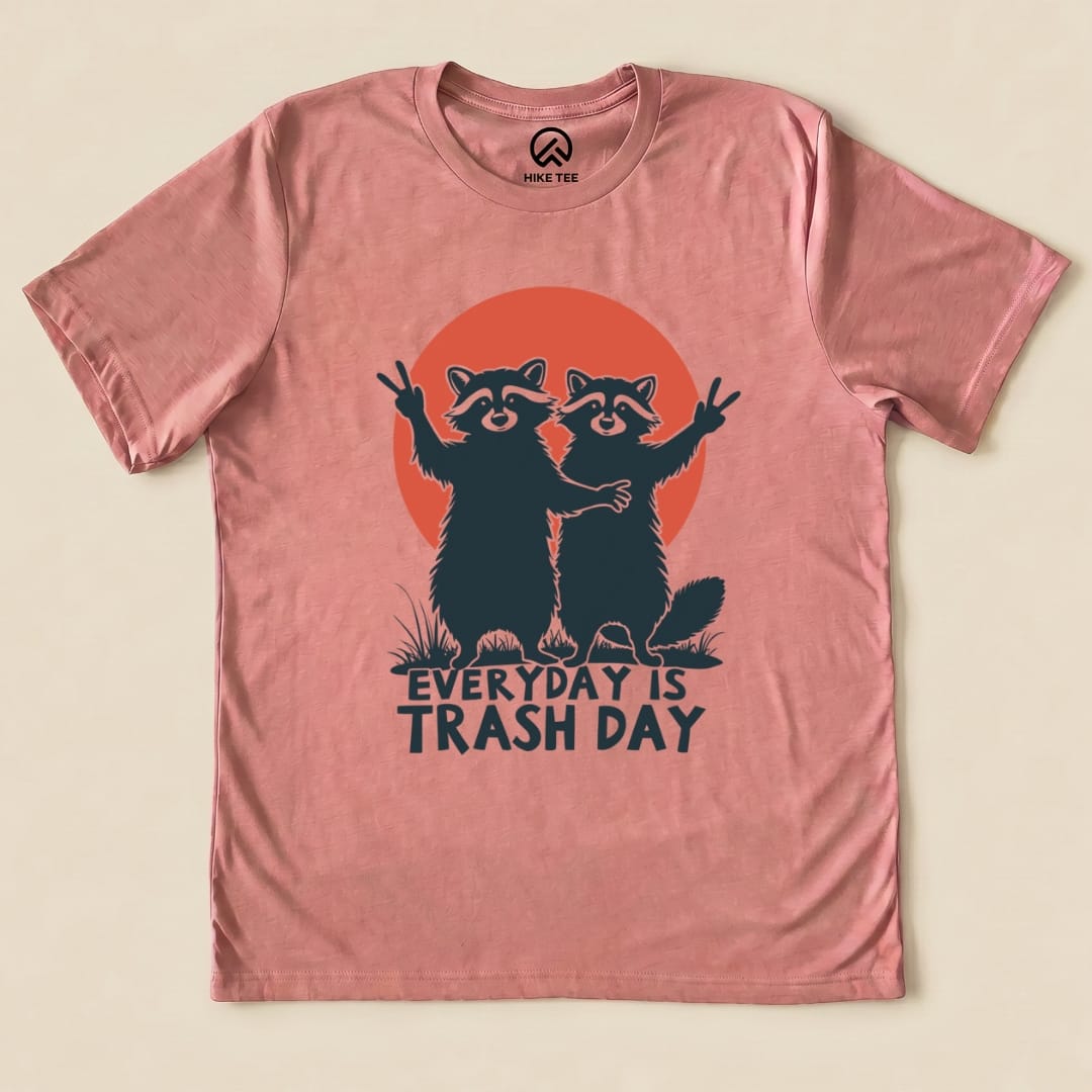 A flat graphic t-shirt in dusty pink featuring two raccoons posing with peace signs and the slogan "Everyday is Trash Day.