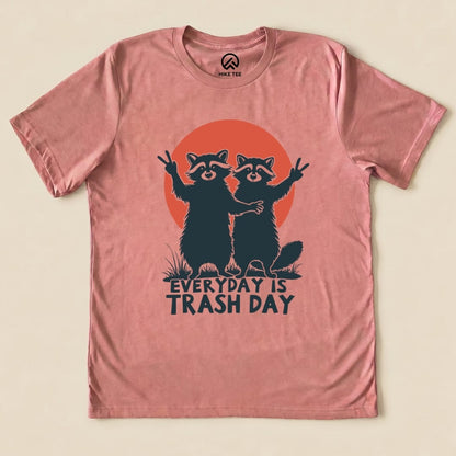 A flat graphic t-shirt in dusty pink featuring two raccoons posing with peace signs and the slogan "Everyday is Trash Day.