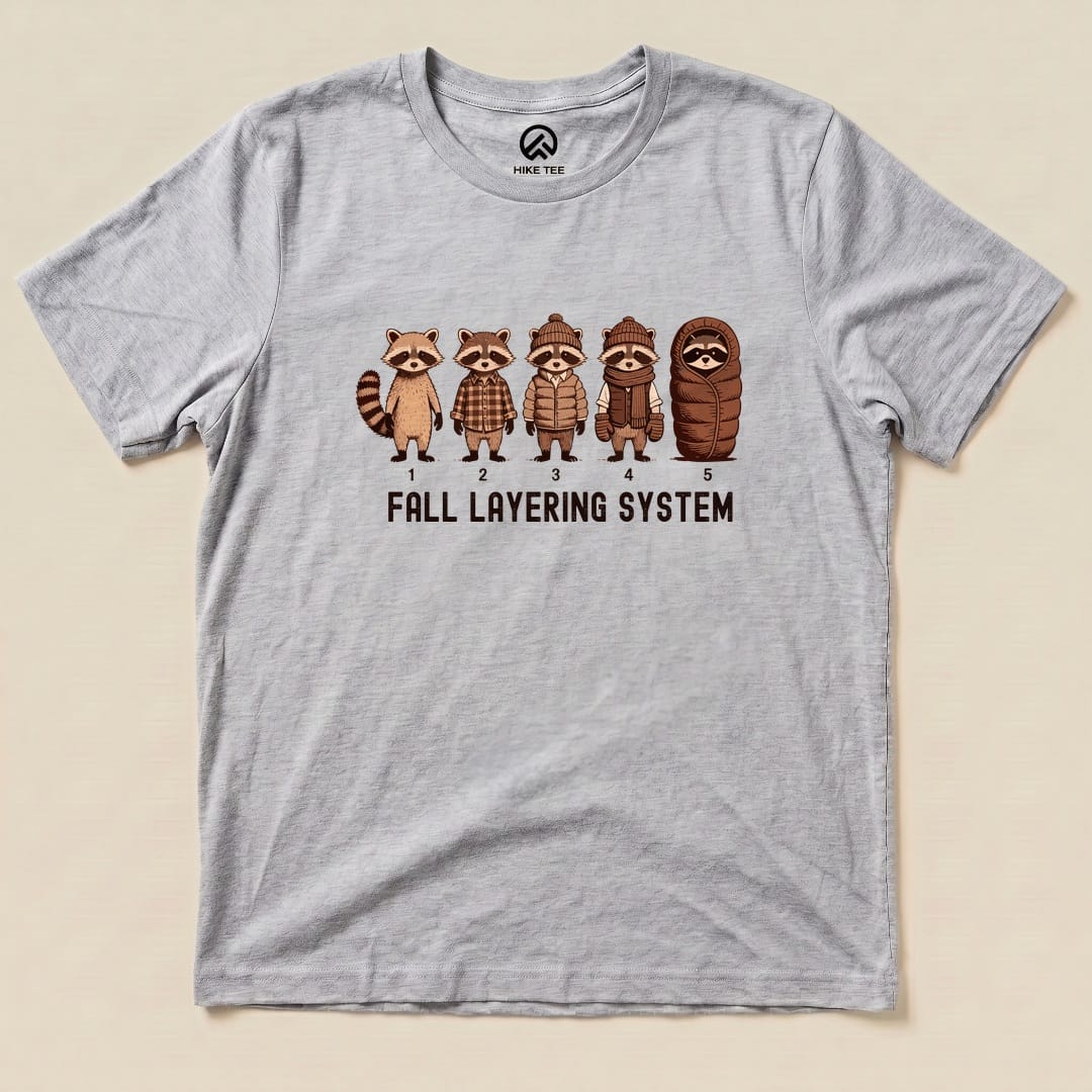A gray graphic t-shirt featuring five cartoon raccoons in various outfits with the words "FALL LAYERING SYSTEM" beneath them.