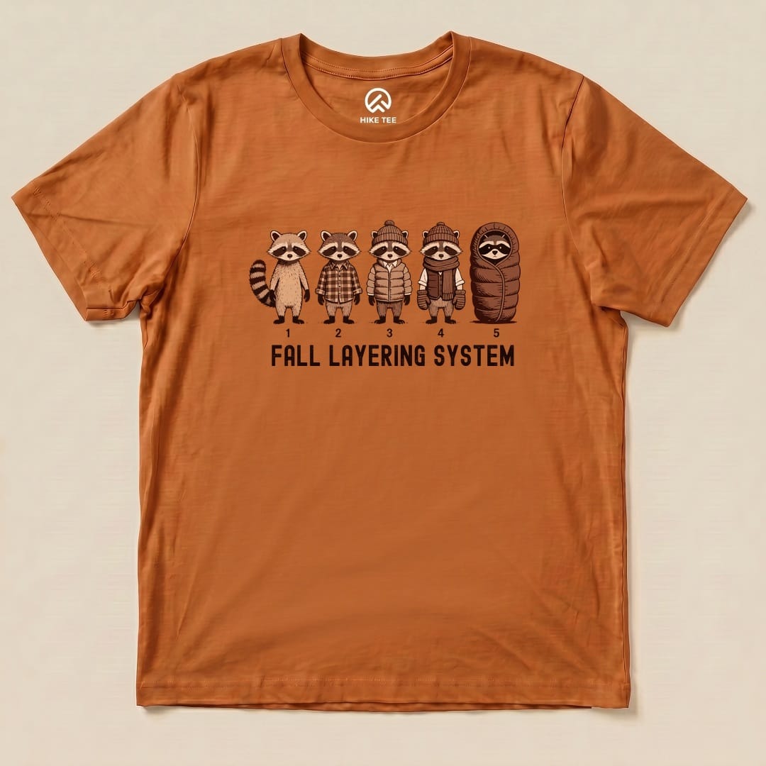 A graphic t-shirt in earthy orange featuring raccoons in various fall outfits with the text "FALL LAYERING SYSTEM.