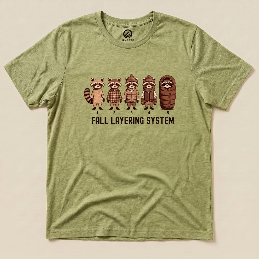A graphic t-shirt featuring five raccoons in different outfits, labeled "Fall Layering System," on an olive green background.