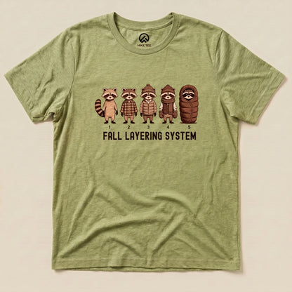 A graphic t-shirt featuring five raccoons in different outfits, labeled "Fall Layering System," on an olive green background.