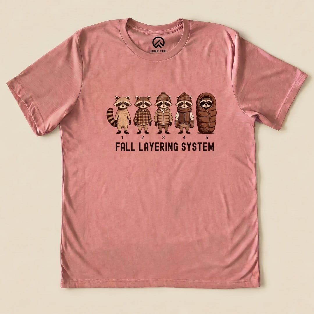 A pink graphic t-shirt featuring raccoons in various fall attire with the text "FALL LAYERING SYSTEM" below them.
