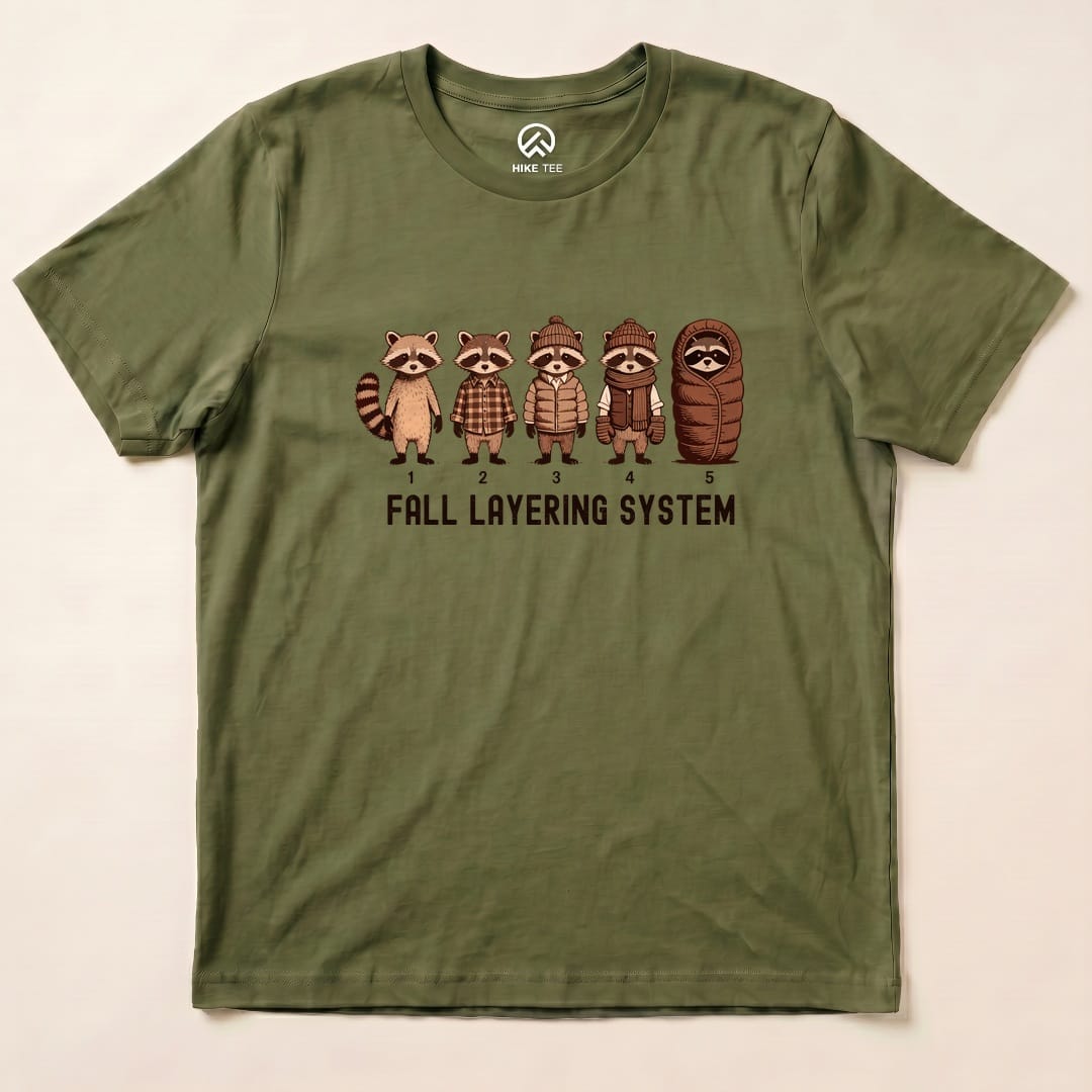 The graphic t-shirt features five raccoons in various clothing layers, with "FALL LAYERING SYSTEM" printed below them.