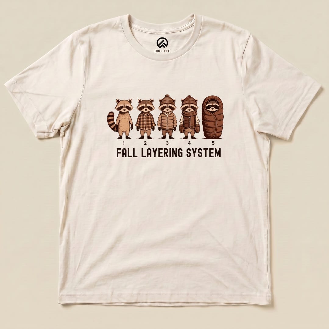 A graphic t-shirt featuring five raccoons depicting the "Fall Layering System" with numbered outfits from casual to cozy.