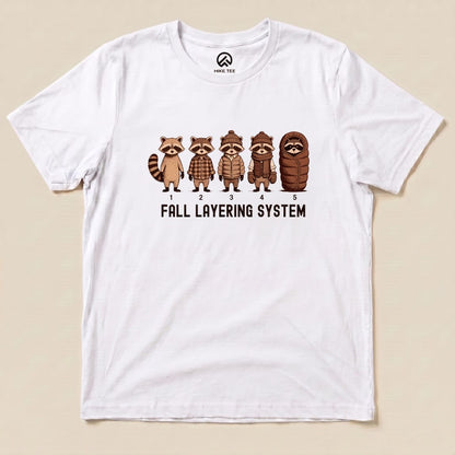 A graphic t-shirt featuring five raccoons in varying outfits with the text "FALL LAYERING SYSTEM" displayed below them.