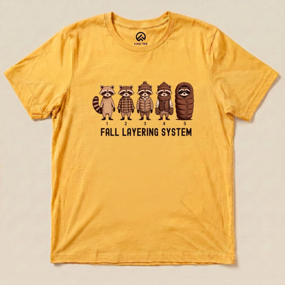 A yellow graphic t-shirt featuring five cartoon raccoons illustrating the "Fall Layering System" with numbered layers.