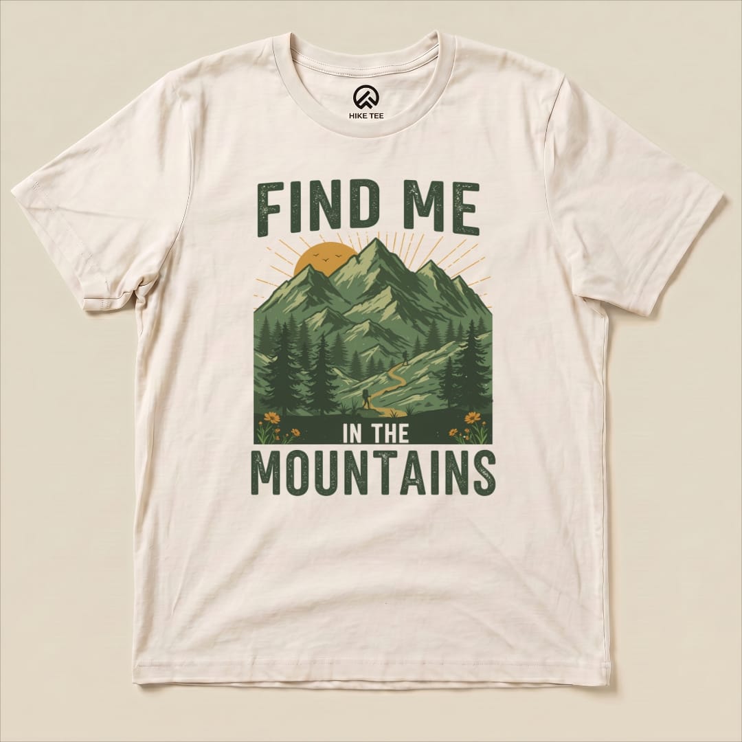 A cream graphic t-shirt featuring the slogan "FIND ME IN THE MOUNTAINS" with a scenic mountain landscape and a winding trail.