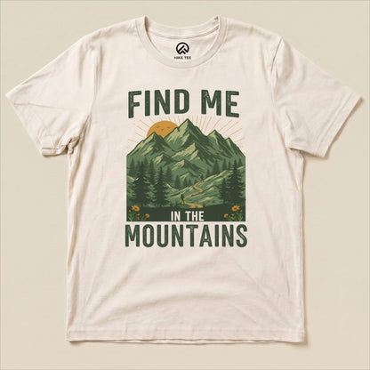 A cream graphic t-shirt featuring the slogan "FIND ME IN THE MOUNTAINS" with a scenic mountain landscape and a winding trail.