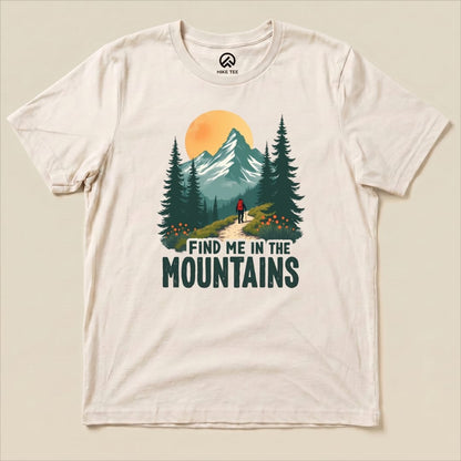 A cream graphic t-shirt featuring a hiker walking on a trail with the text "FIND ME IN THE MOUNTAINS" and vibrant mountain scenery.