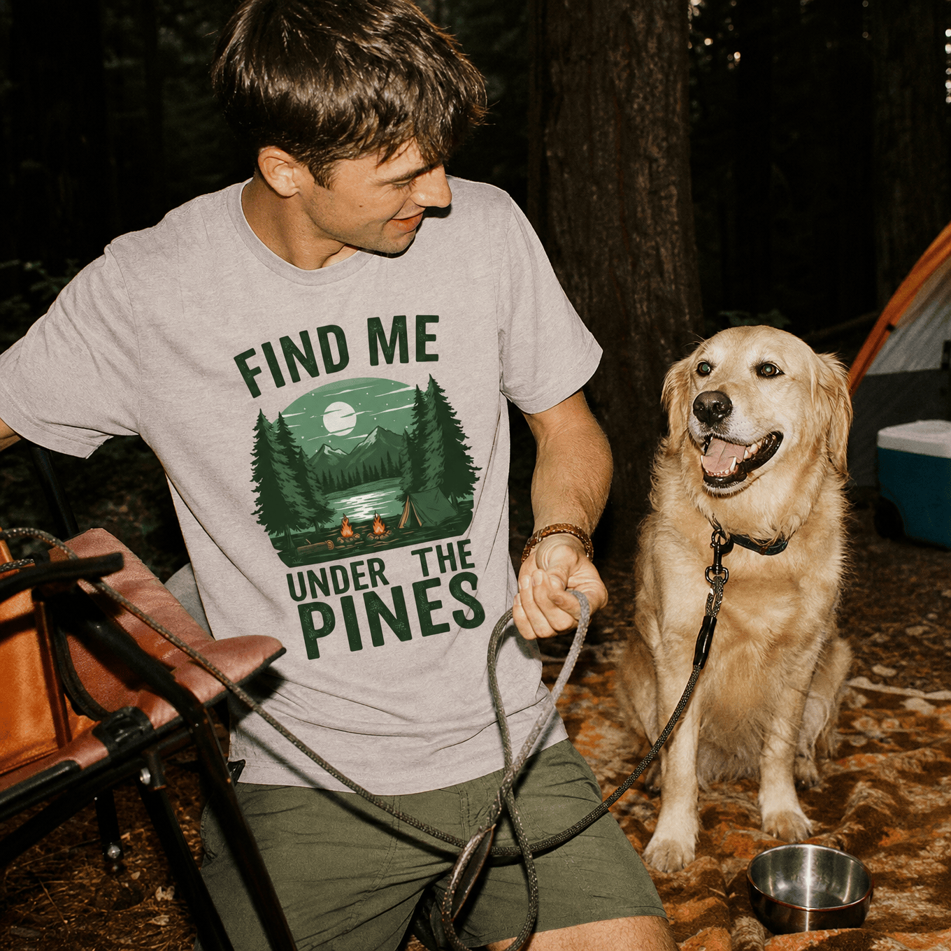 A man sits in a forest wearing a graphic t-shirt that reads "Find Me Under The Pines," with a golden retriever nearby.