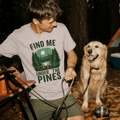 A man sits in a forest wearing a graphic t-shirt that reads "Find Me Under The Pines," with a golden retriever nearby.