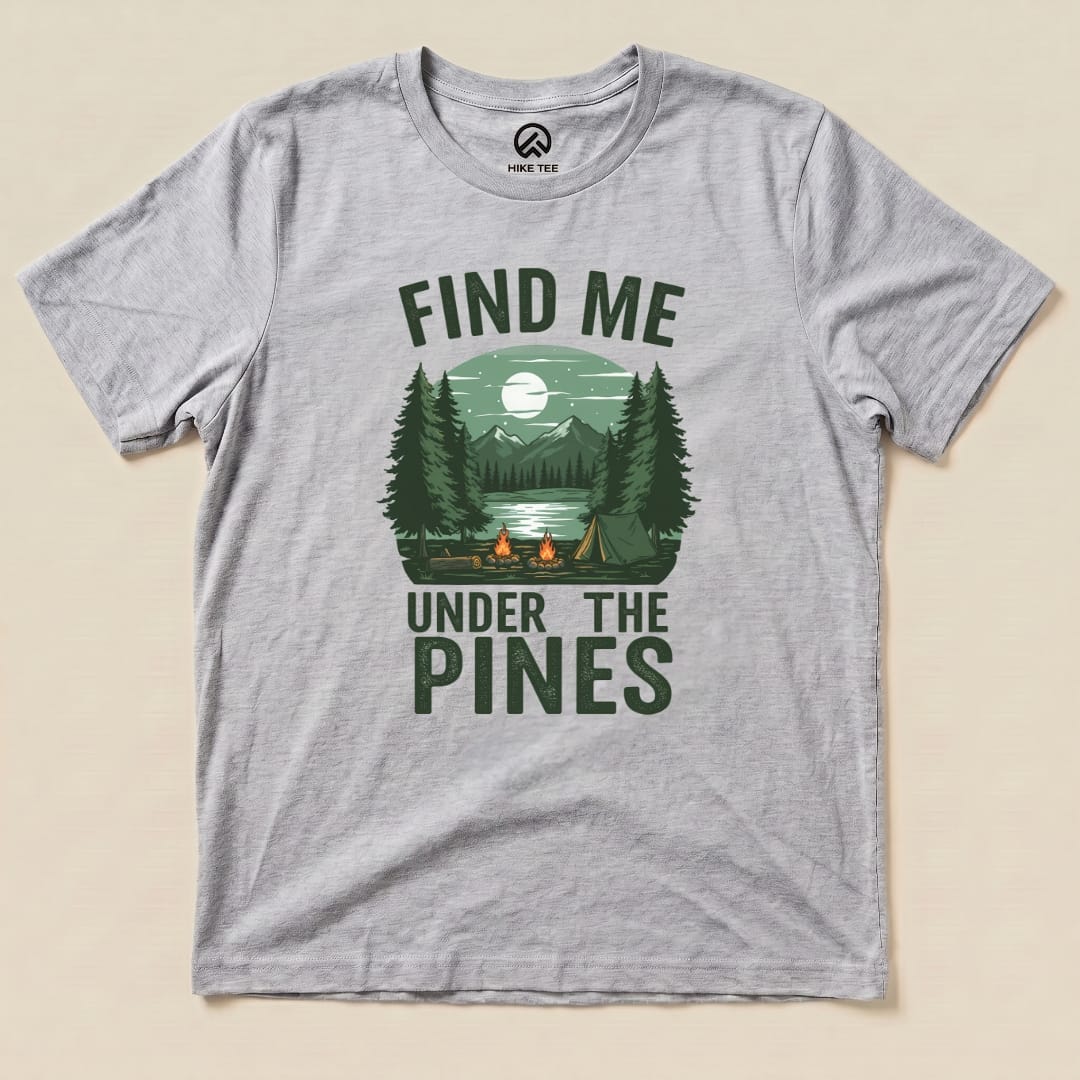 A gray graphic t-shirt features a serene camp scene with the words "Find Me Under The Pines" above a lake and mountains.