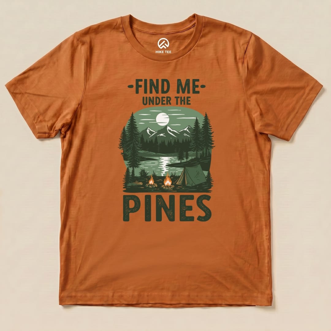 The orange graphic t-shirt features an outdoor scene with a lake, mountains, and pine trees, topped by the phrase "Find Me Under The Pines Shirt.