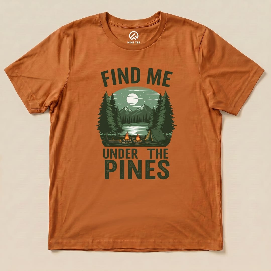 The "Find Me Under The Pines Shirt" features a serene camping scene with trees, a lake, and a mountain backdrop in earthy tones.