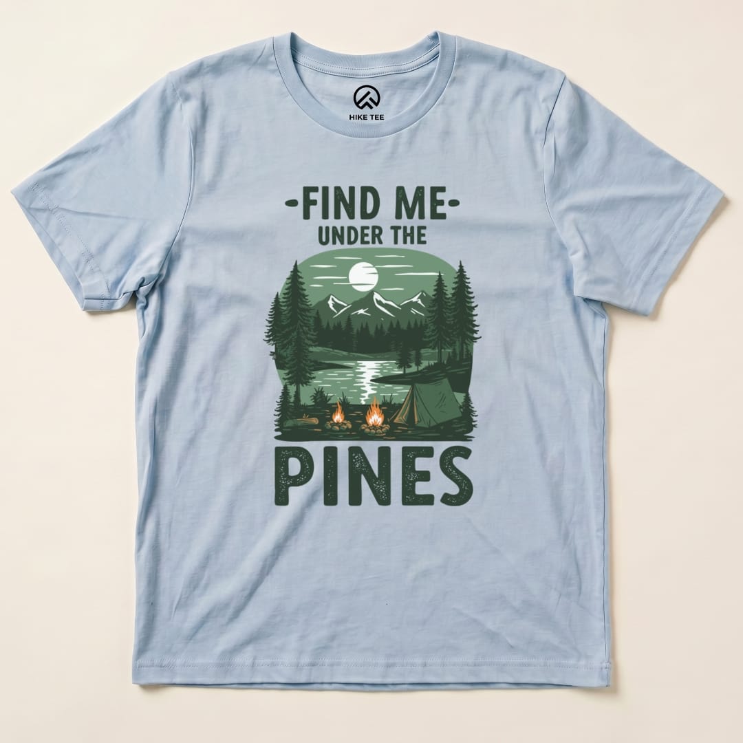 A light blue graphic t-shirt featuring the phrase "Find Me Under The Pines" along with a scenic illustration of a campsite, trees, and mountains.