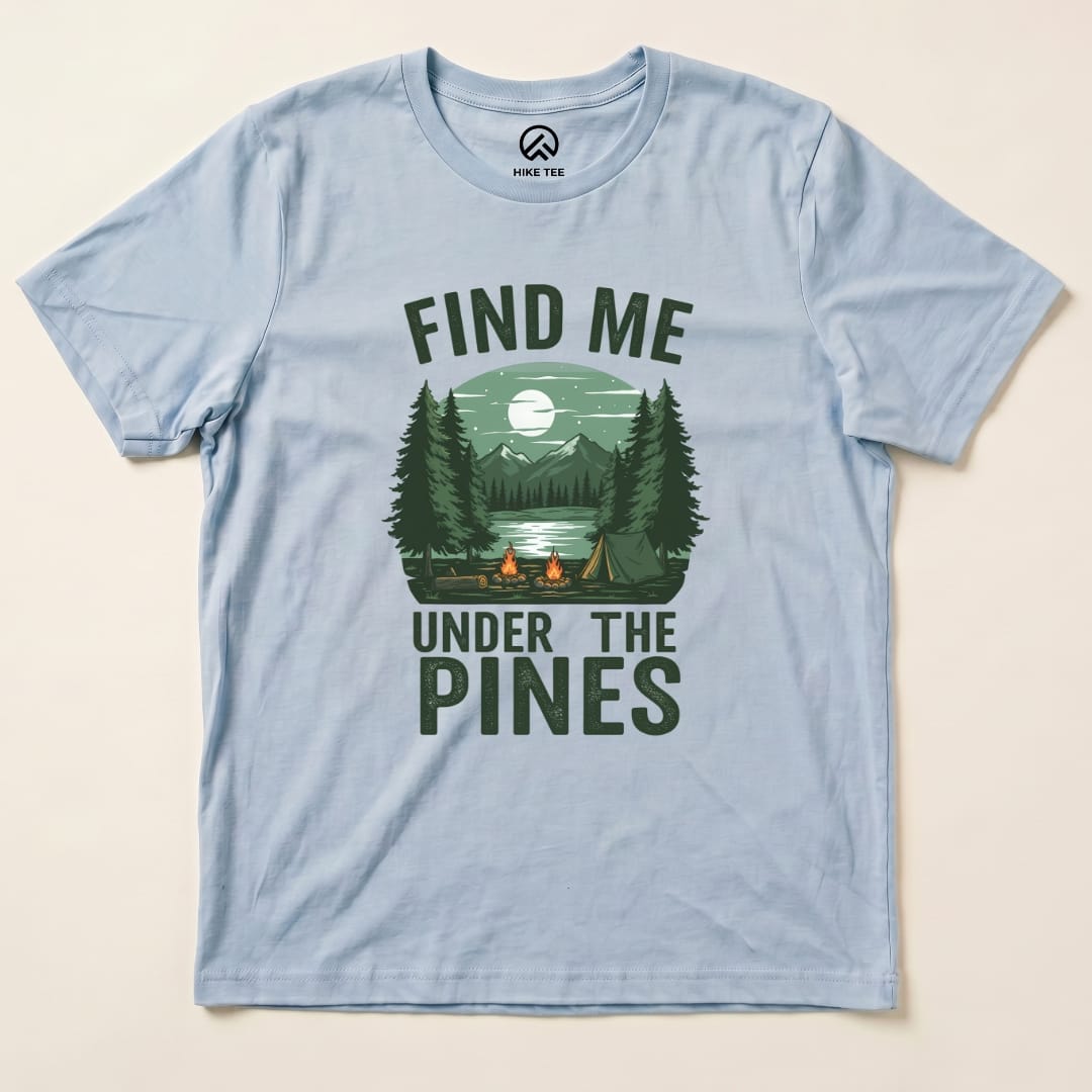 A light blue graphic t-shirt featuring a campfire scene with trees and mountains, reading "Find Me Under The Pines Shirt.