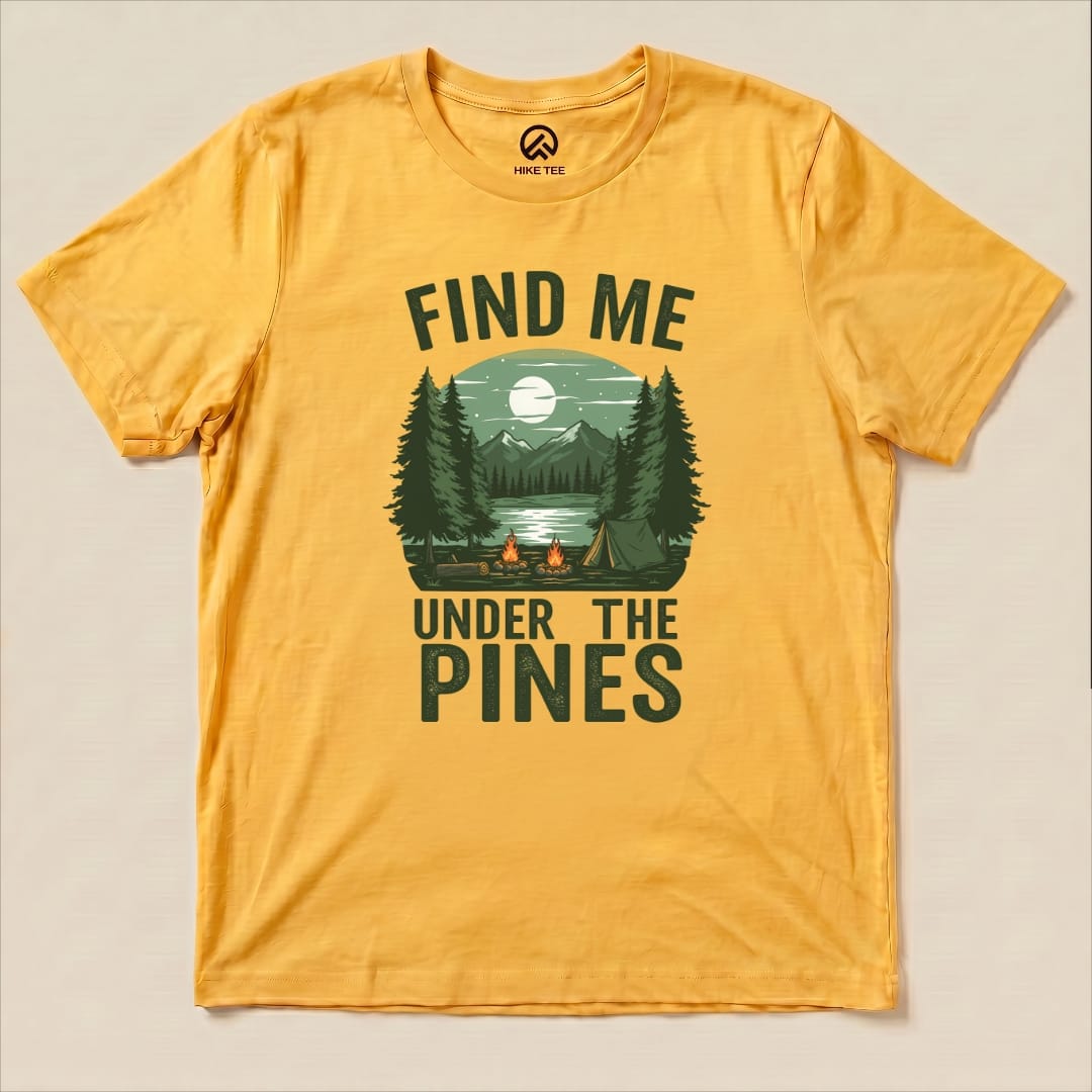 Graphic t-shirt featuring the phrase "Find Me Under The Pines" with a serene forest scene, mountains, and a campfire design.