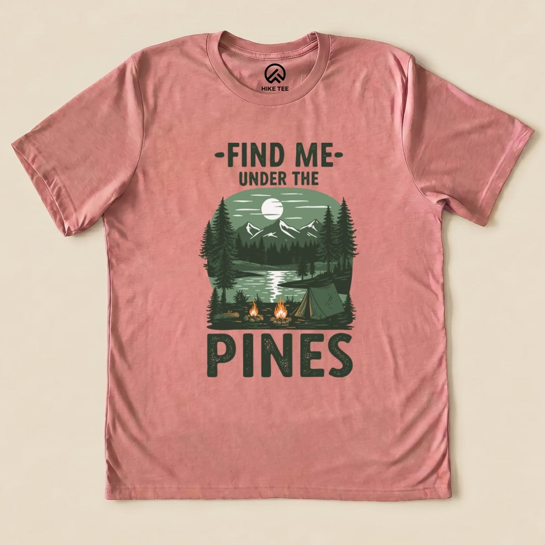 A graphic t-shirt in pink featuring the phrase "Find Me Under The Pines" with a scenic forest and campsite design.