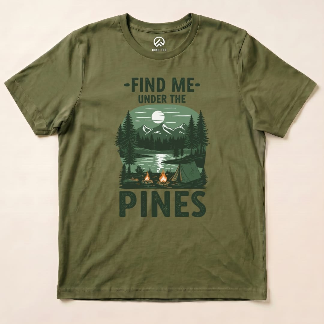 The olive green graphic t-shirt features a scenic design with the words "Find Me Under The Pines" above a forest and campsite.