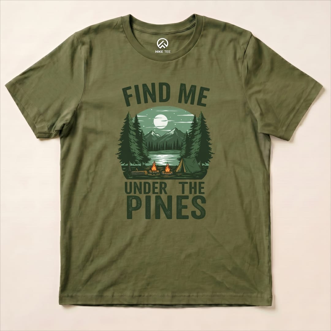 A flat lay of the green "Find Me Under The Pines Shirt" featuring a graphic of a serene camping scene with mountains, trees, and a lake.