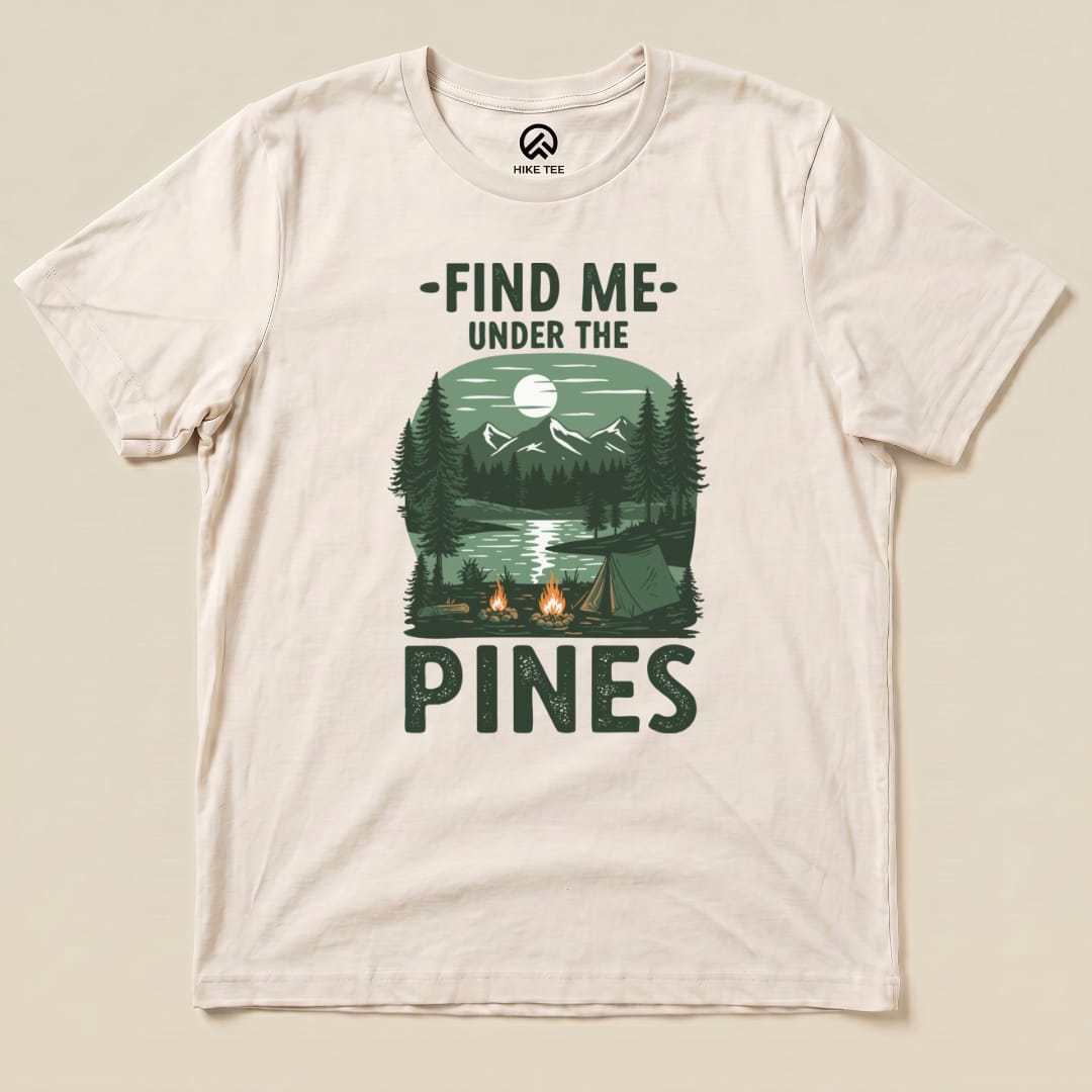 A graphic t-shirt featuring the slogan "Find Me Under The Pines" and a scenic design of mountains, a lake, and campfires.