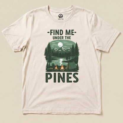 A graphic t-shirt featuring the slogan "Find Me Under The Pines" and a scenic design of mountains, a lake, and campfires.