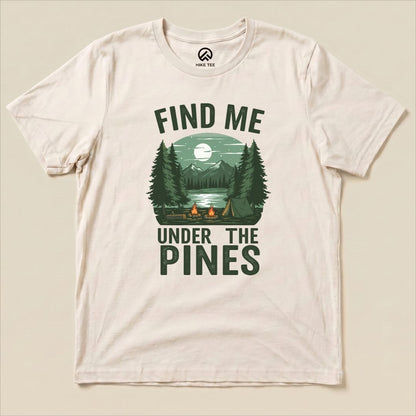 A flat graphic t-shirt featuring the design "Find Me Under The Pines," with a scenic campfire, mountains, and pine trees illustration.