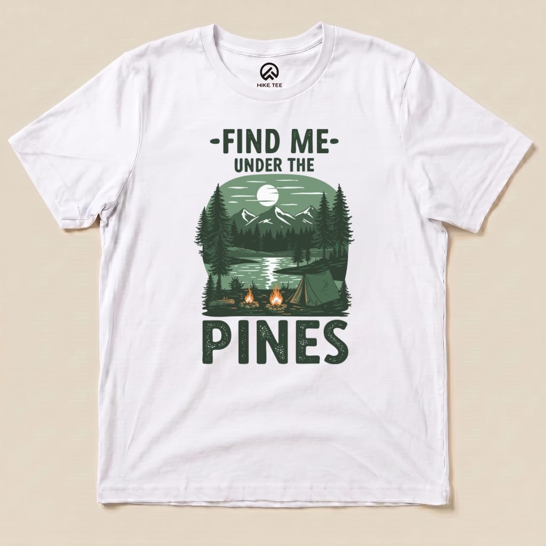 Graphic t-shirt featuring a serene forest landscape with the slogan "Find Me Under The Pines" and imagery of mountains, trees, and a lake.