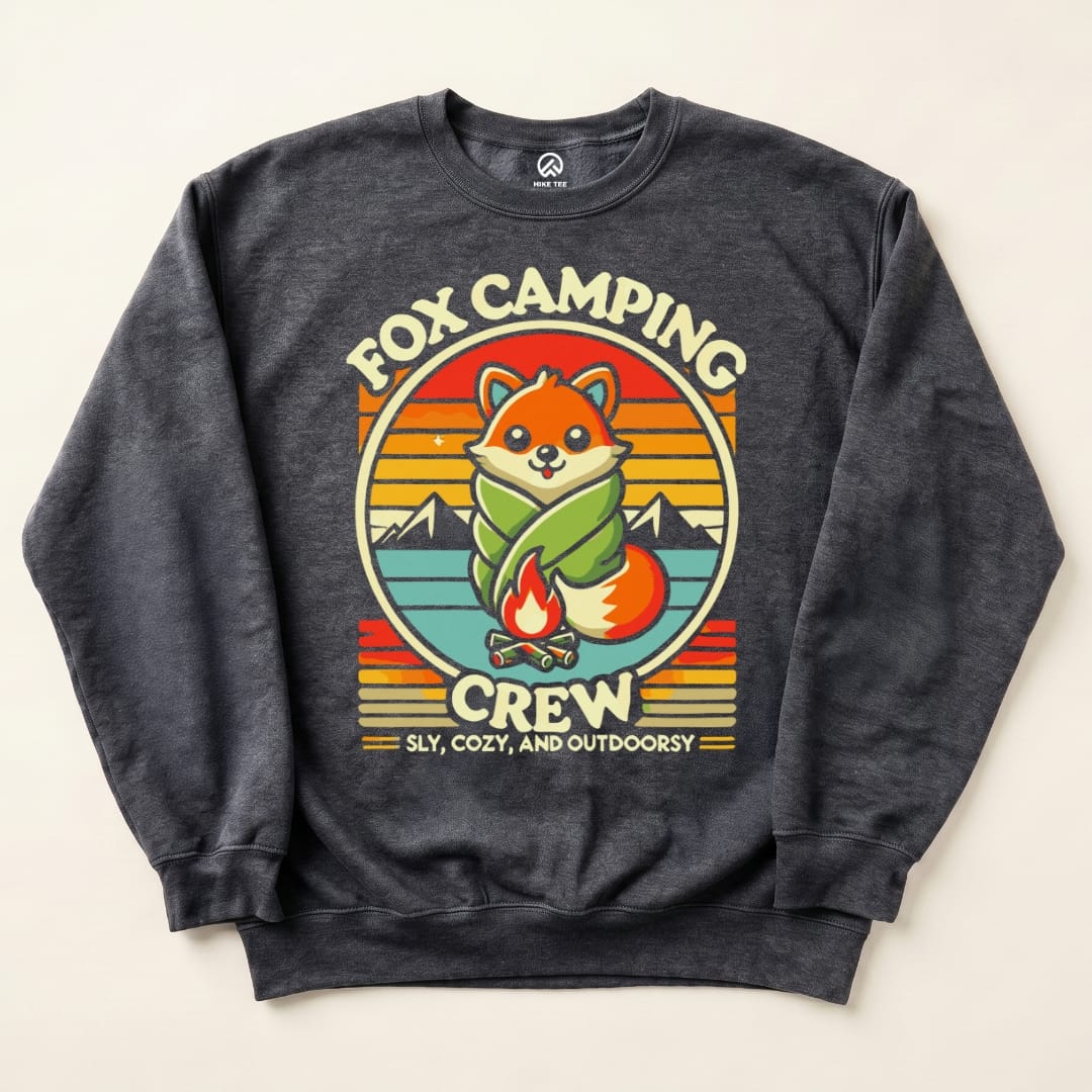A graphic sweatshirt featuring a cute fox wrapped in a sleeping bag, surrounded by mountains, with the text "Fox Camping Crew.