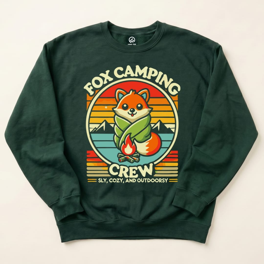 A dark green Fox Camping Crew Sweatshirt features a playful fox wrapped in a blanket by a campfire with mountains in the background.