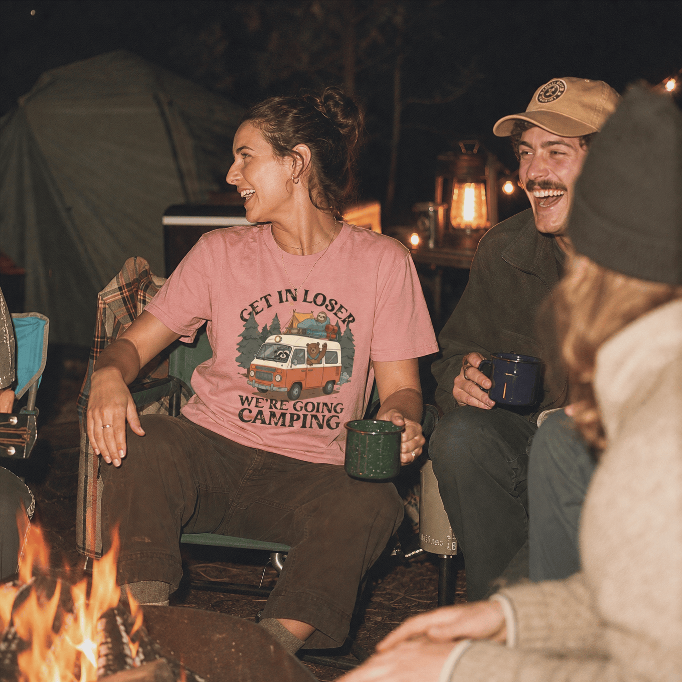 A woman sitting by a campfire wears a pink graphic t-shirt that reads "Get In Loser We're Going Camping," featuring a vintage camper and trees.