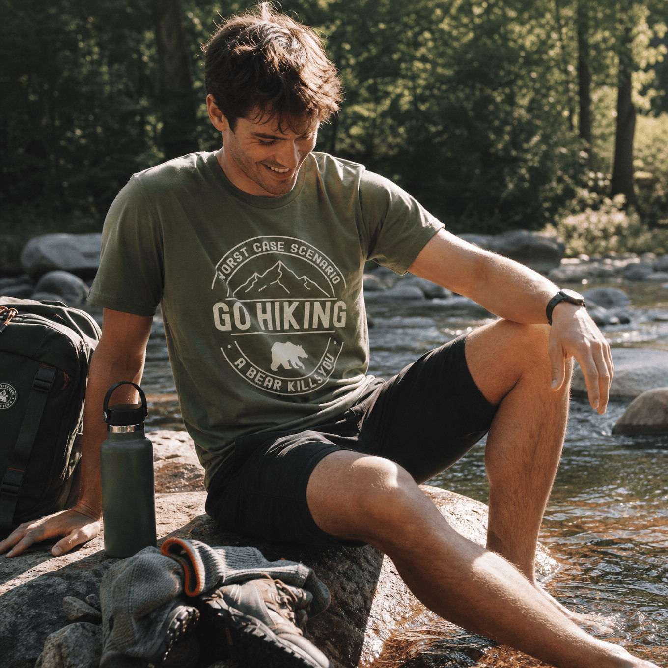 A man sitting on a rock by a stream wears a graphic t-shirt featuring the slogan "Go Hiking Worst Case Scenario A Bear Kills You.