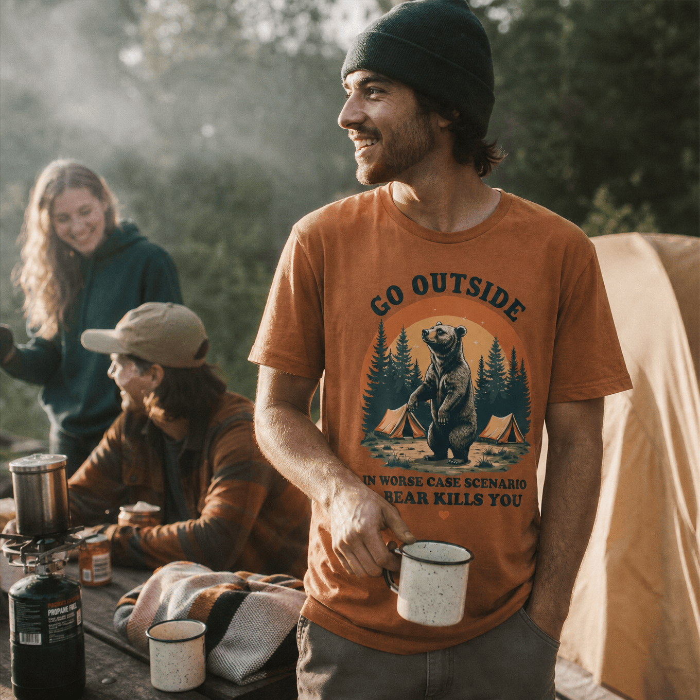 A man in a campsite smiles while holding a mug, wearing a graphic t-shirt featuring a bear and the words "Go Outside Shirt.