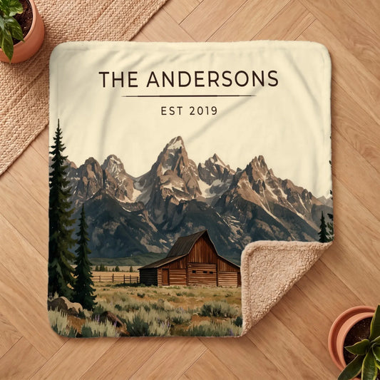 A cozy Grand Teton Barn Sherpa Blanket featuring a rustic barn and towering mountains with "THE ANDERSONS EST 2019" text.