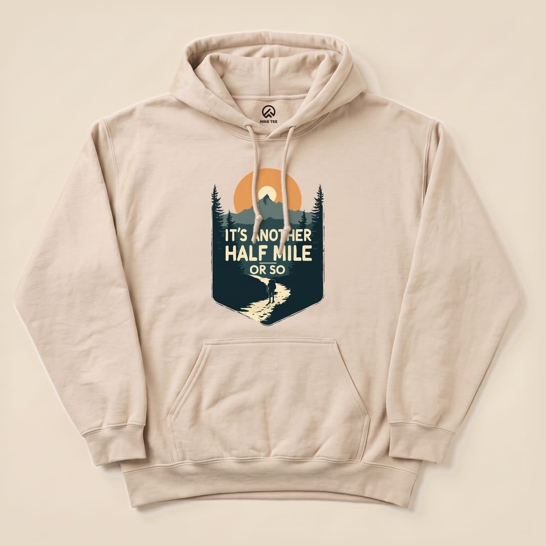 A beige Half Mile Hoodie featuring a graphic of a hiker on a trail beneath the sun, with the text "IT'S ANOTHER HALF MILE OR SO.