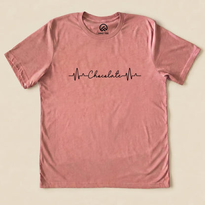 A pink graphic t-shirt featuring the phrase "Chocolate" with heartbeat lines, labeled as the Heart Beat Chocolate Shirt.