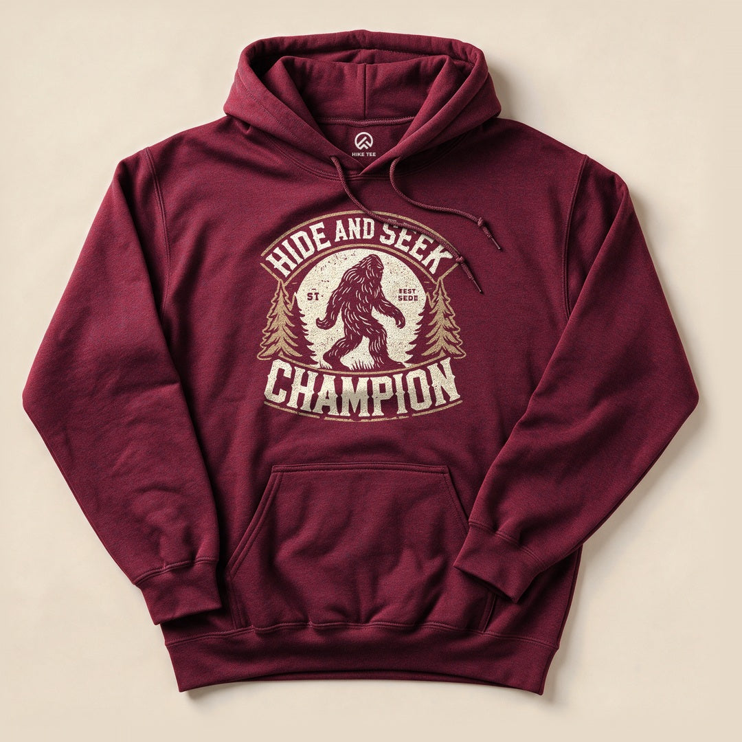 A maroon hooded sweatshirt featuring a graphic of Bigfoot with the phrase "Hide and Seek Champion" above and trees on the sides.