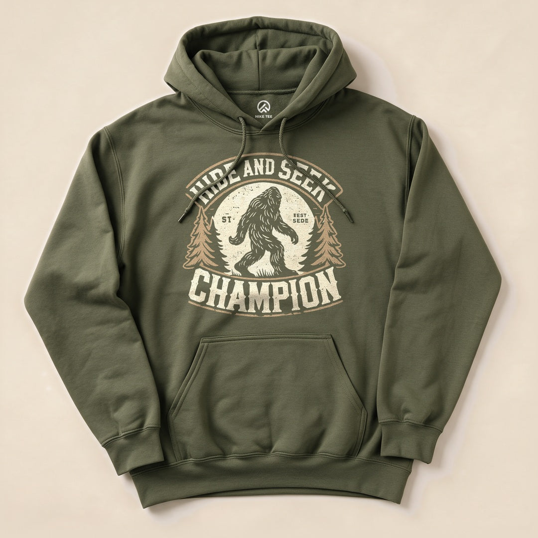 The olive green Hide and Seek Champion Bigfoot Hoodie features a graphic of Bigfoot with trees and bold text in a vintage style.
