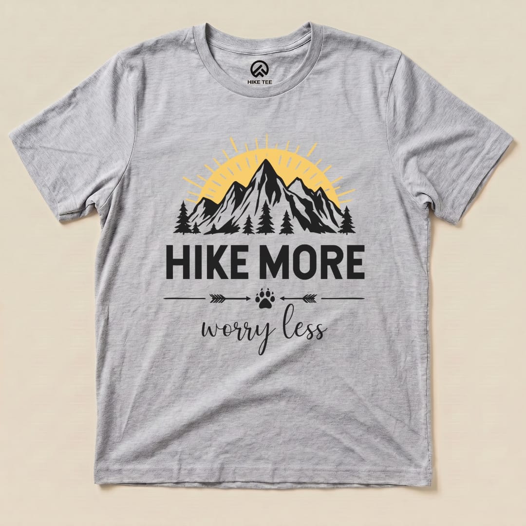 A flat lay of a gray graphic t-shirt featuring mountains, trees, and the slogan "Hike More Worry Less" in bold black letters.