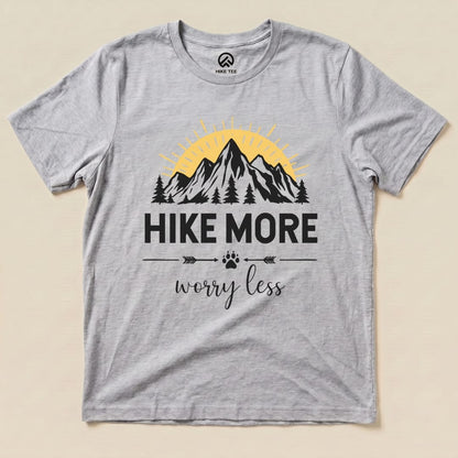 A flat lay of a gray graphic t-shirt featuring mountains, trees, and the slogan "Hike More Worry Less" in bold black letters.