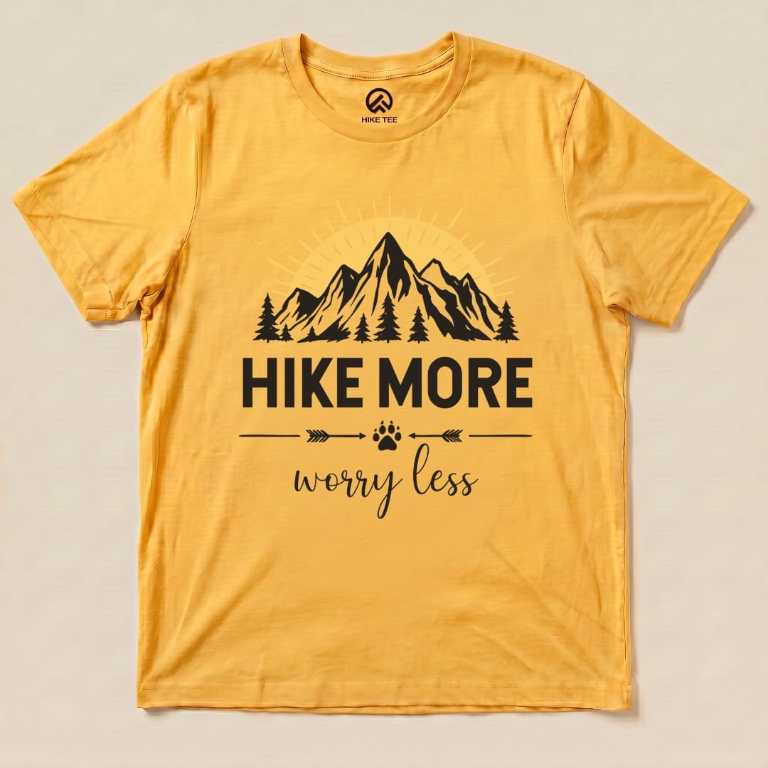 A flat graphic t-shirt in yellow featuring the slogan “Hike More Worry Less” along with mountain and tree illustrations.