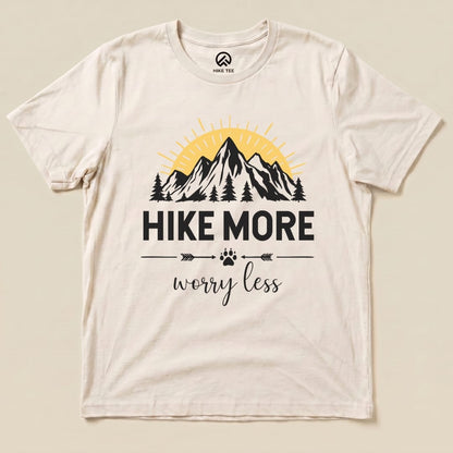 A flat lay of a beige graphic t-shirt featuring the slogan "Hike More Worry Less" with mountain and sun designs printed on it.